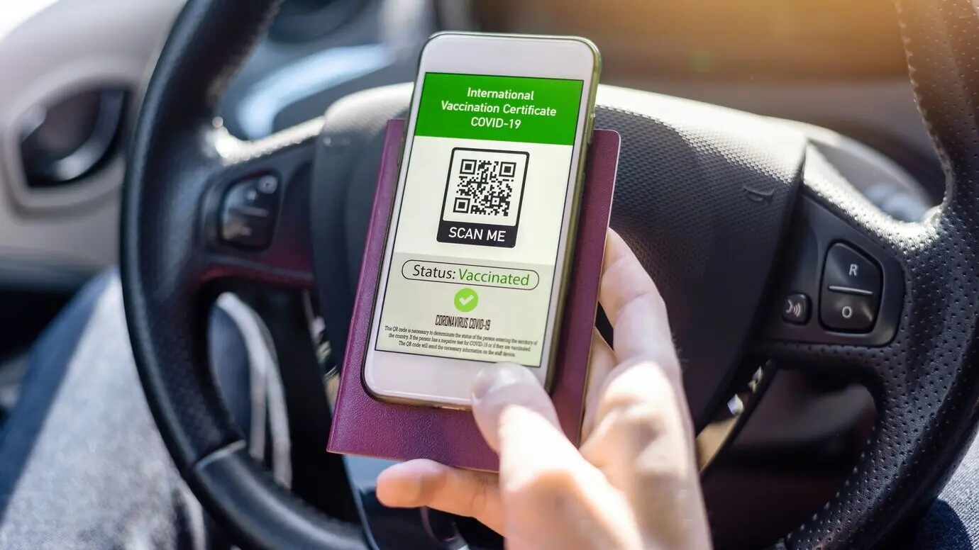 Eine männliche Hand hält im Auto einen Reisepass und ein Smartphone mit dem QR-Code des internationalen COVID-19-Impfzertifikats.
